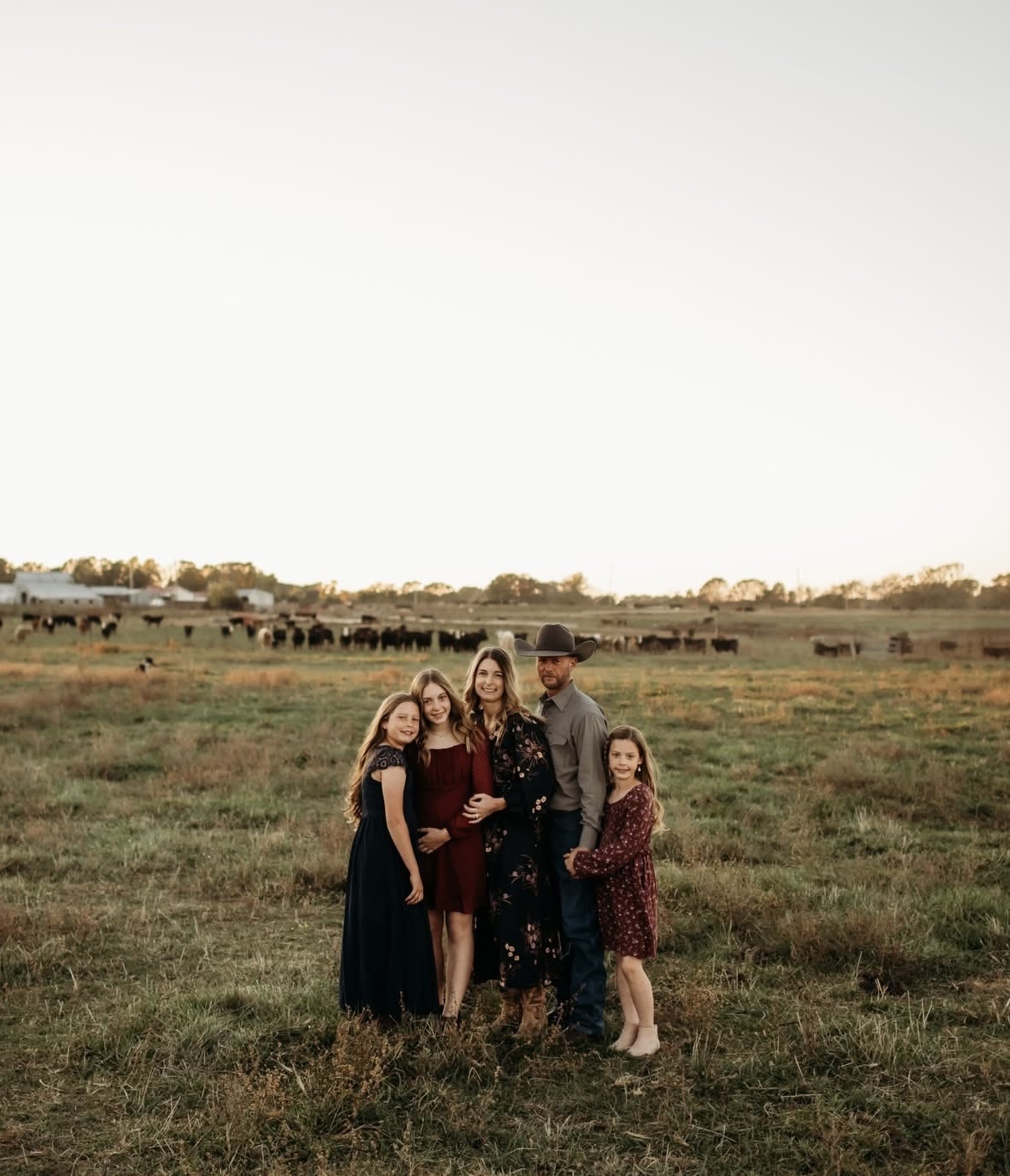 Family of 3 girls, wife, and man with a cowboy hat standing in a field with cattle in the background