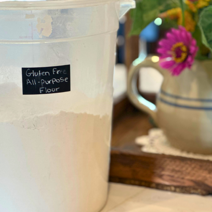 all purpose gluten free flour in a container with a label and a pitcher of flowers next to it